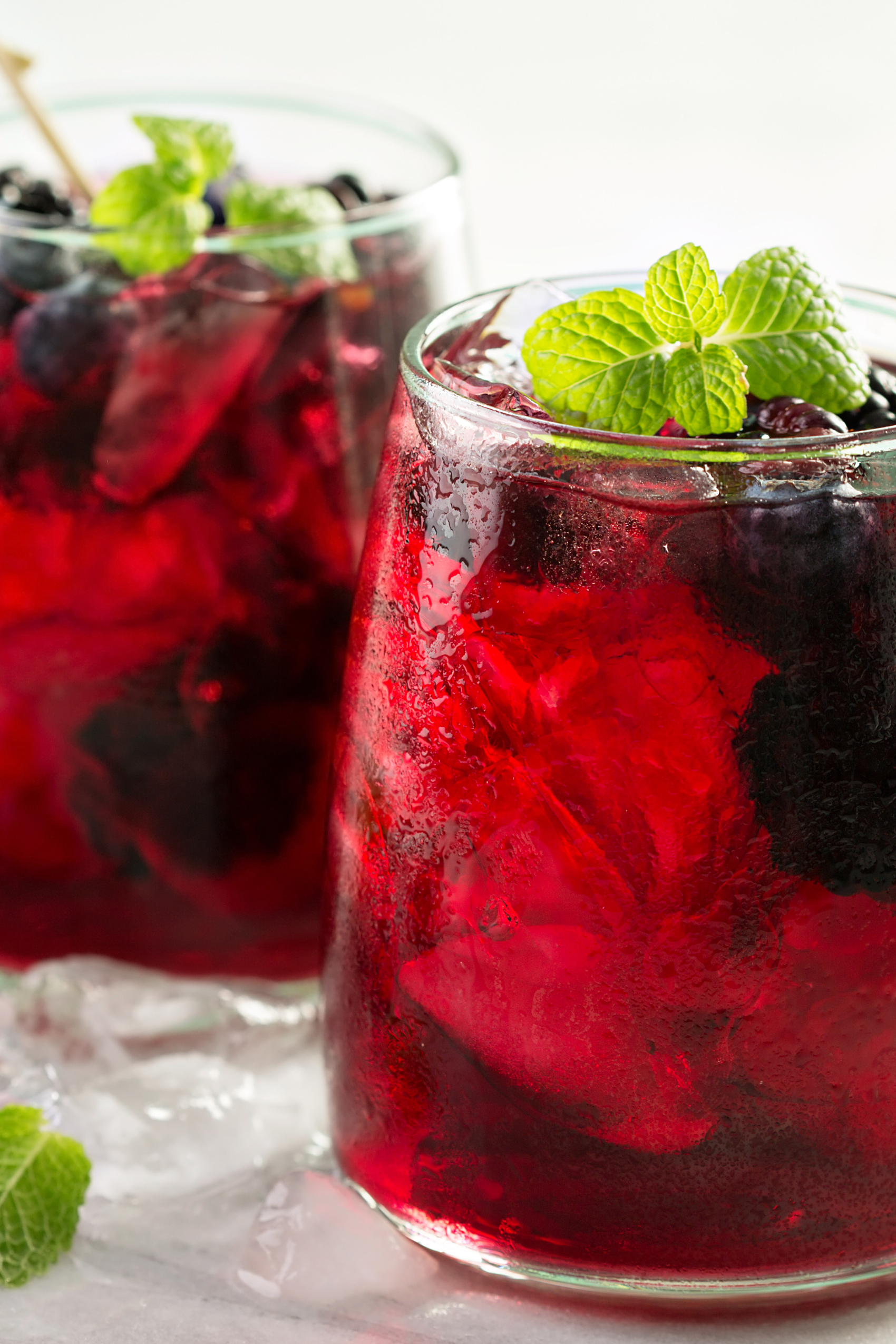 Two glass cups back left and front right filled with a bright red cold drink with berries topped with mint leaves.