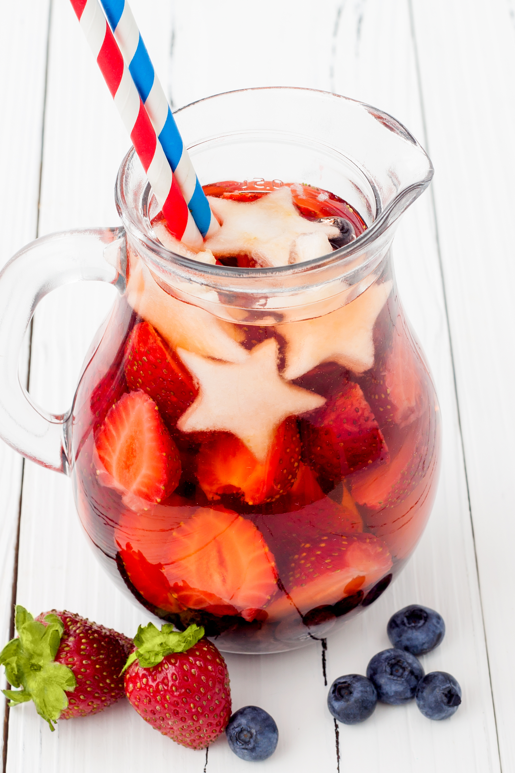 A glass pitcher filled with sliced strawberries and star shaped apples topped with two straws on a white wooden background with strawberries and blueberries placed in front of pitcher.